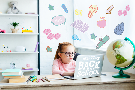 Beautiful child in eyeglasses using laptop while studying at desk at home welcome back to school Stock Photo