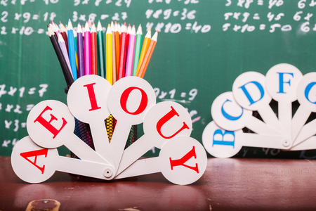 Colorful pencils of red yellow orange violet purple pink green and blue in stationary cup ruler and fan english alphabet standing on brown school desk on written with white chalk blackboard on math Stock Photo