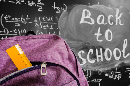 Back to school background with purple school bag with yellow ruler and the title back to school and math formulas written by white chalk on the black school chalkboard